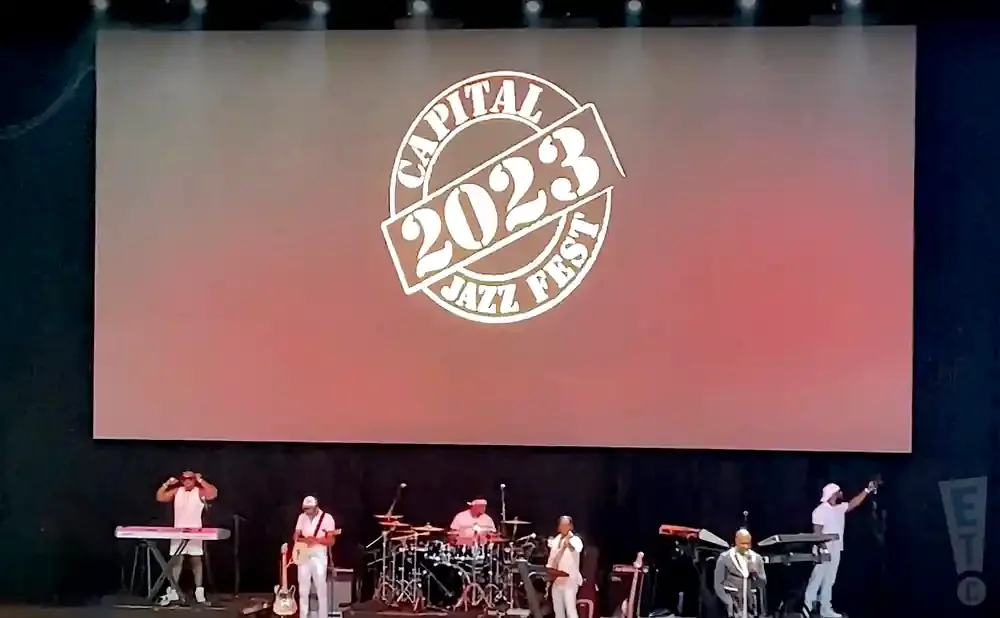 A band performs on stage in front of a large screen displaying the 