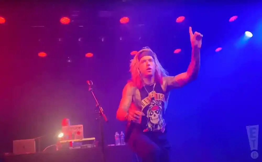 A male performer with long blonde hair and a bandana sings into a microphone on a stage with red and blue lights.