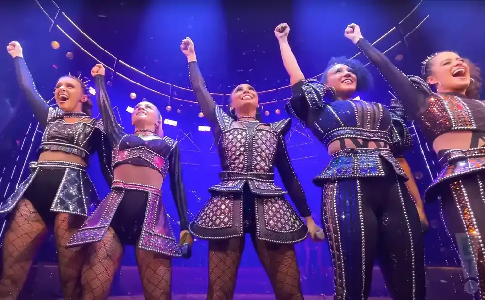 Five women in sparkly black and jewel-toned costumes, with raised fists, perform on a stage with a blue background and gold circular lights.