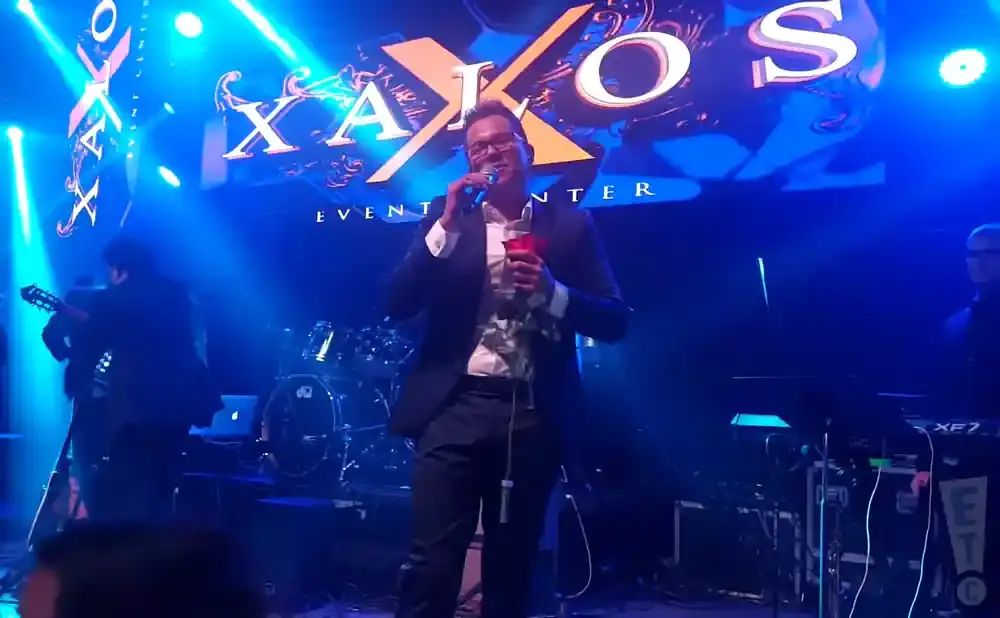 A singer in a suit holds a rose and microphone on stage with a band and a 