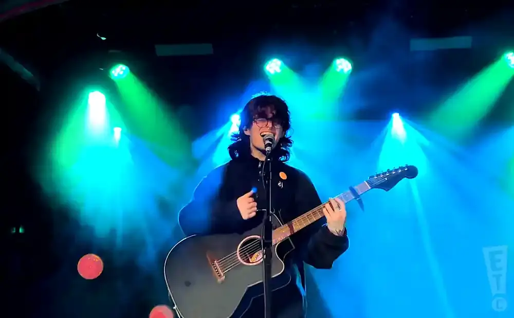 A person with dark hair and glasses sings and plays an acoustic guitar on a stage lit with blue and green lights.