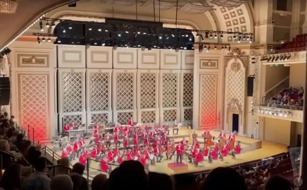 An orchestra performs in a grand concert hall with intricate white wall paneling and an audience in the foreground.