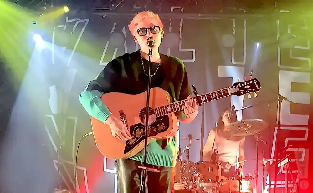 A musician with pink hair plays an acoustic guitar on stage, with a drummer in the background.