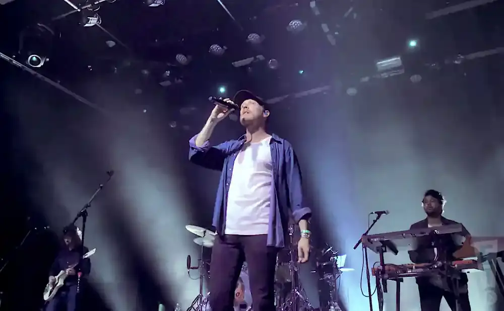 A singer in a blue shirt and white t-shirt holds a microphone on stage with a band.