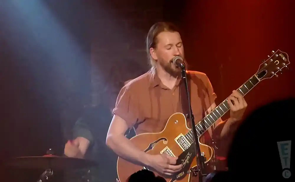 A man with long hair plays a brown guitar and sings into a microphone on a dimly lit stage.