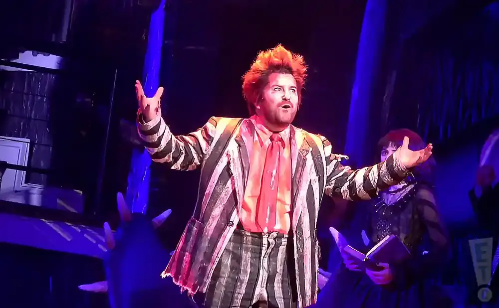 A man with spiky orange hair in a striped suit and tie, on stage with other performers.