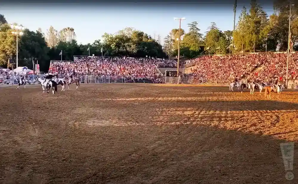 A rodeo crowd watches cowboys on horseback in a dirt arena during a sunset.
