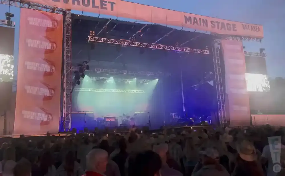 A large outdoor stage at a music festival is lit with spotlights and fog machines, with a banner reading 