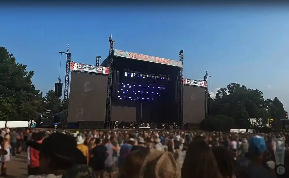 Outdoor concert stage with large screens and a crowd of people. 