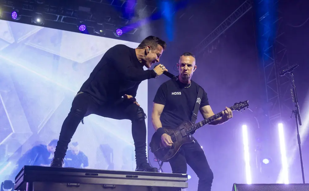 A male singer and guitarist perform on a brightly lit stage with purple lights and a large screen.