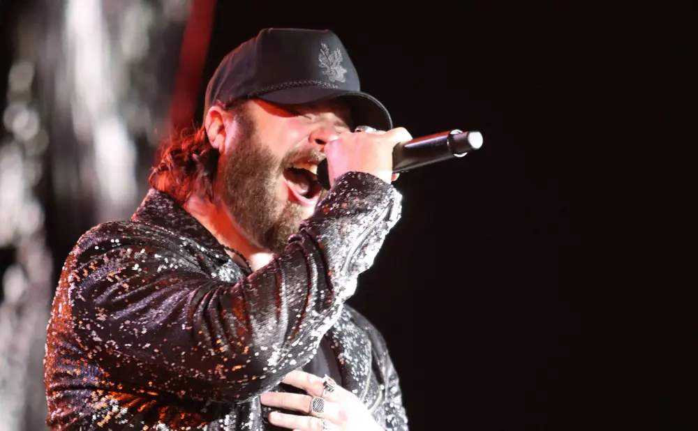 A man with a beard sings into a microphone, wearing a sparkly jacket and a black baseball cap.