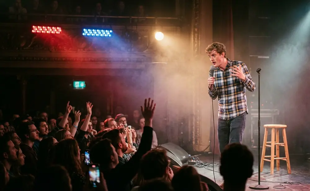 A comedian performs on stage with a microphone, while the audience cheers with hands raised.