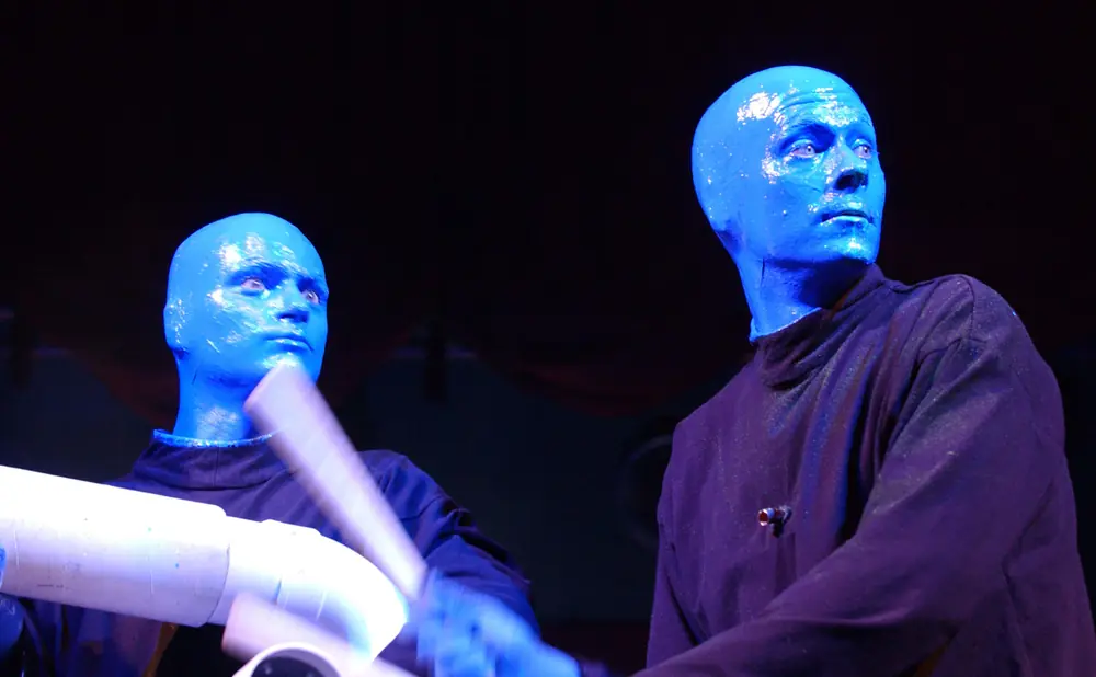 Blue Man Group performers, one with arms blurred in motion playing on white tubes.