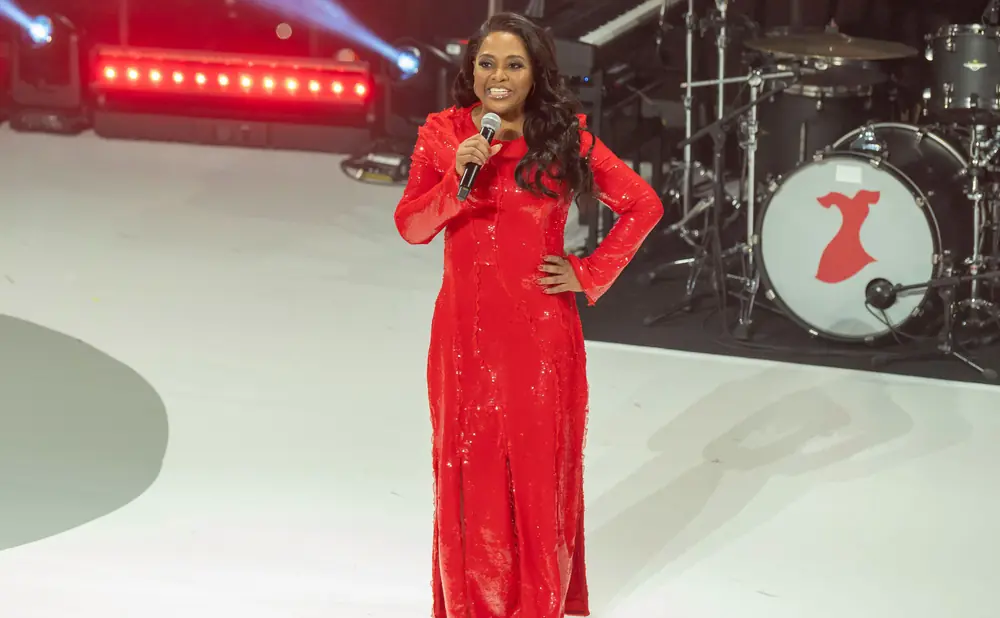 A woman in a sparkling red dress speaks into a microphone on a stage with drums in the background.