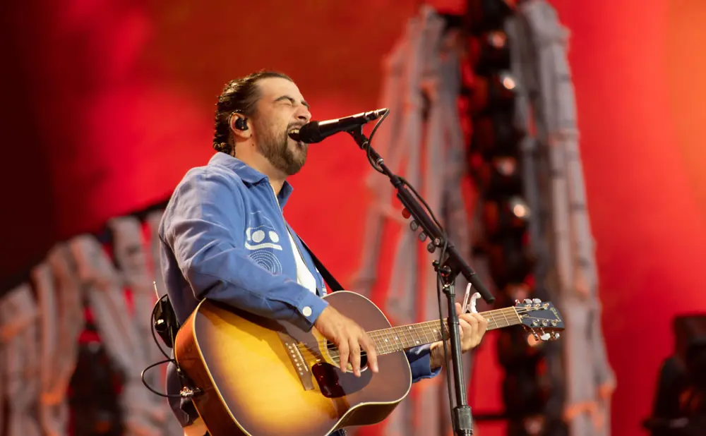 A man with a beard sings into a microphone while playing an acoustic guitar on stage.