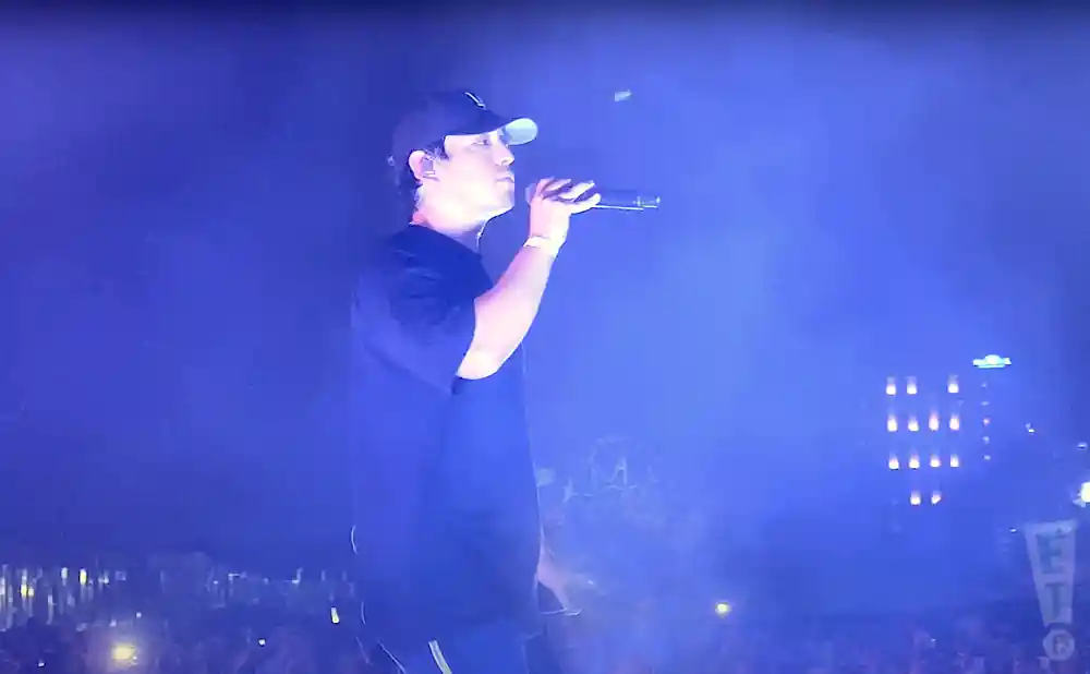 A man in a black t-shirt and baseball cap sings into a microphone on a brightly lit stage.