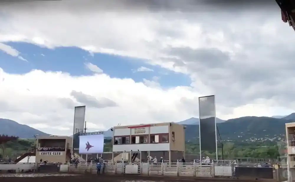 A rodeo arena with a scoreboard showing a jet, tall mirrored structures, and a crowd under a cloudy sky.