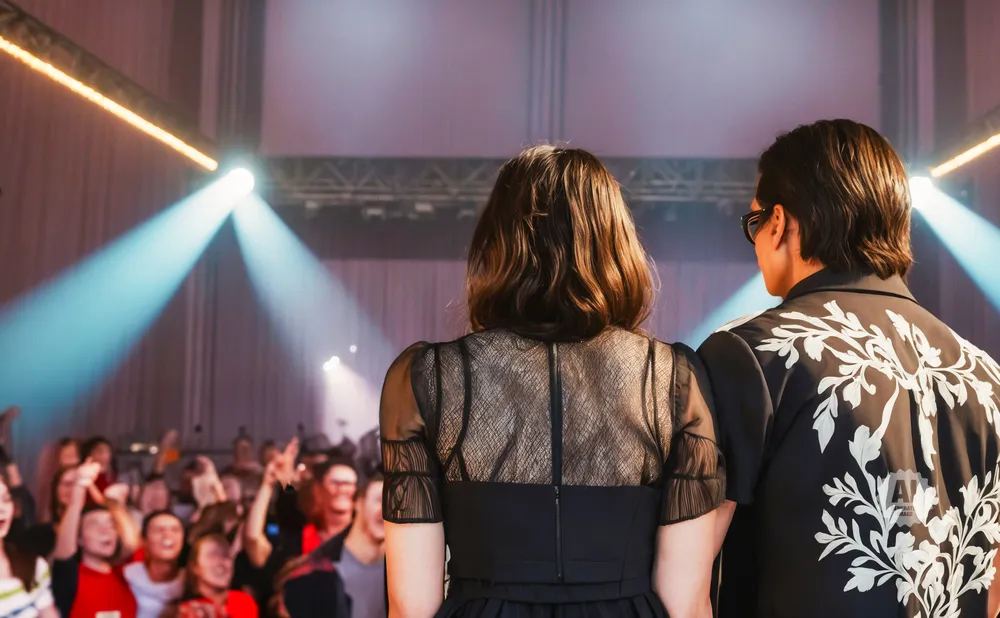 Two people face away from the camera on a stage, with a crowd and stage lights in the background.