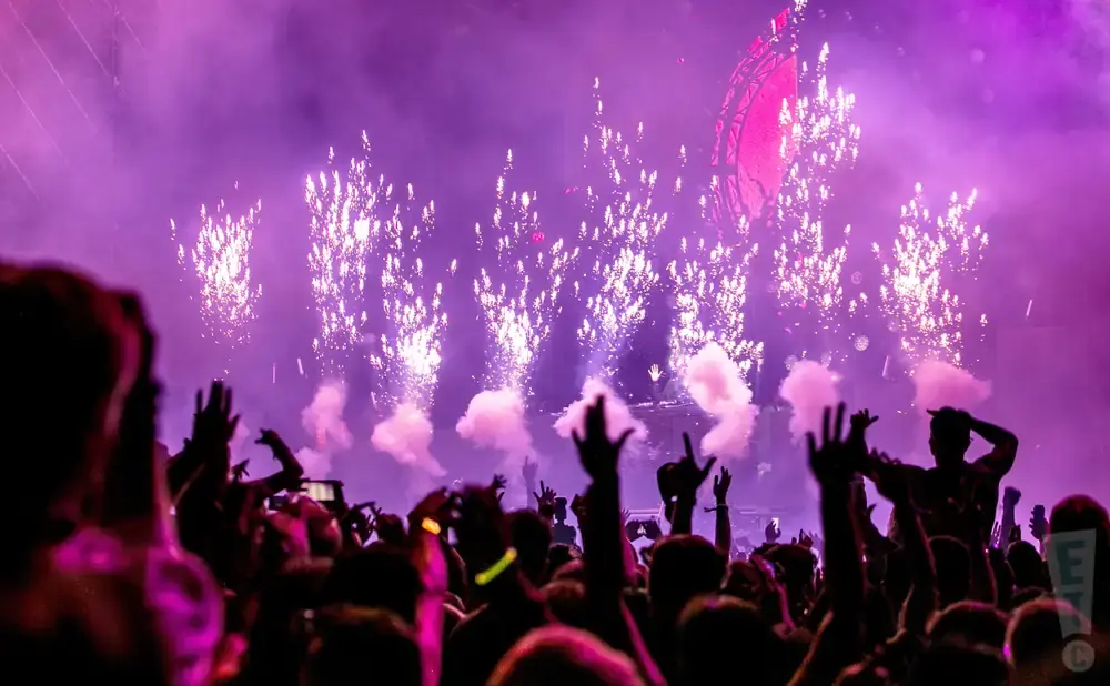 A crowd cheers at a concert with purple fireworks and smoke.