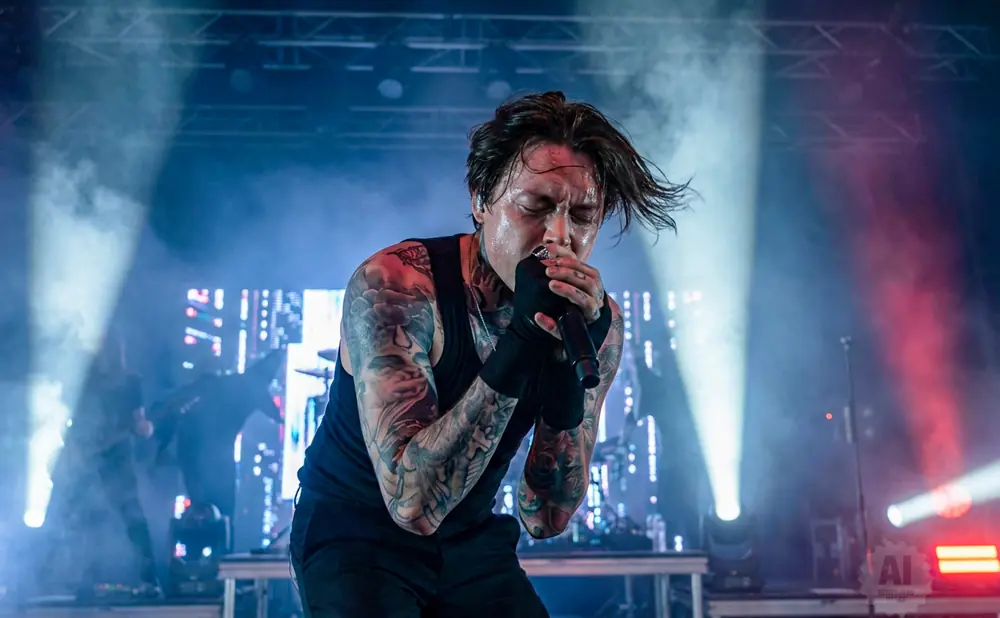 A male singer with tattoos on his arms and neck sings passionately into a microphone on a smoky stage with bright lights.
