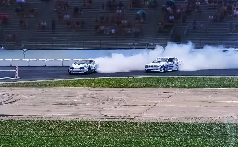 Two white drift cars with blue flames are creating large smoke plumes on a race track, with spectators in the background.