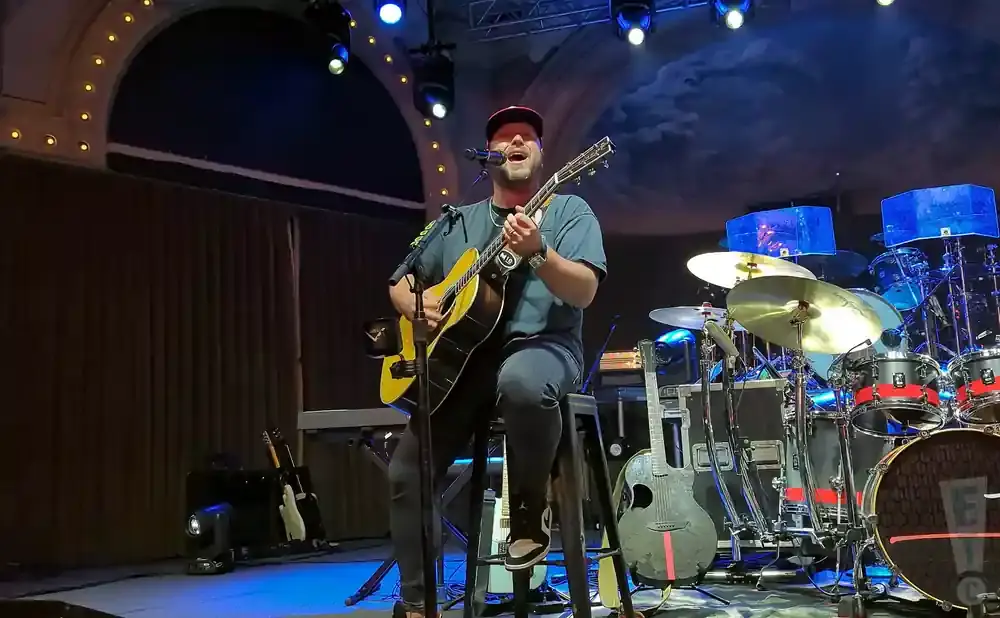 A man in a red hat plays an acoustic guitar while sitting on a stool on a stage with drums and other instruments.