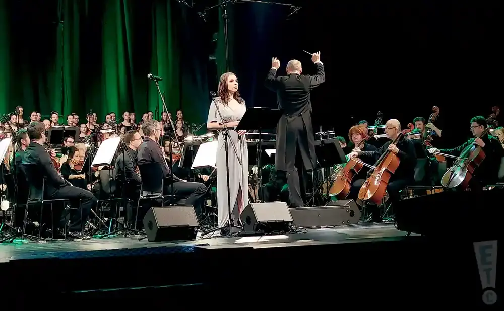 A female singer and conductor lead an orchestra and choir on a stage with green backdrops.