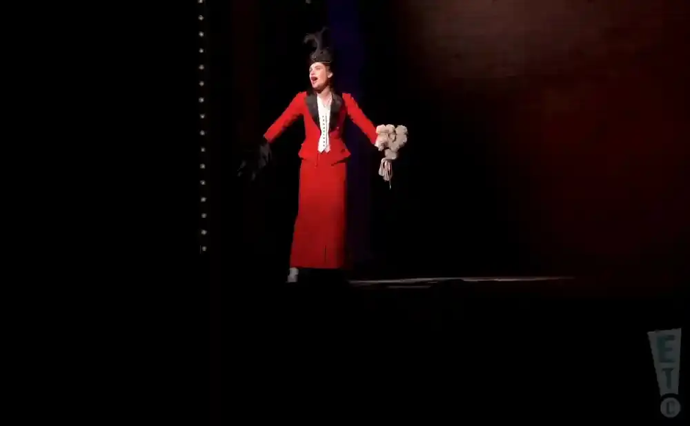 A performer in a red suit and hat sings on stage, holding a bouquet of white flowers.