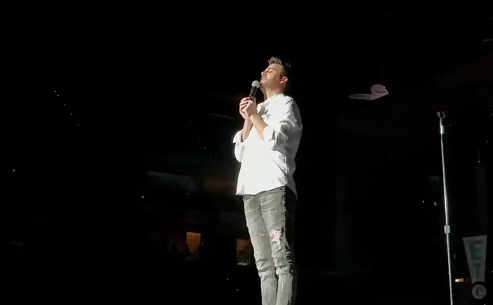 A man in a white shirt and ripped jeans sings into a microphone on a dark stage.