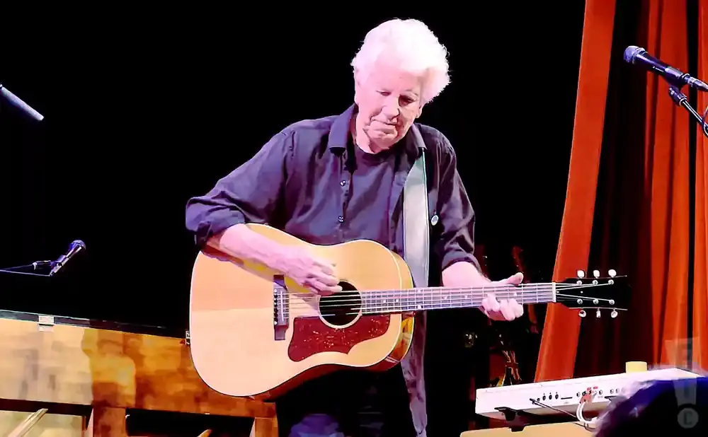 Musician with white hair plays an acoustic guitar on stage.