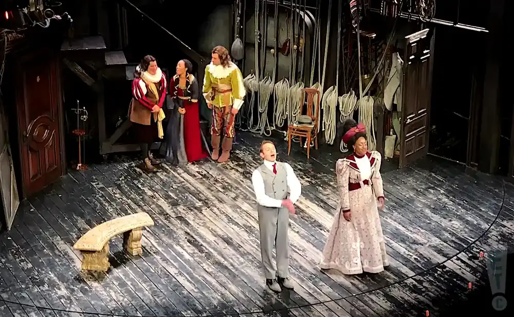 A group of actors in costume on a wooden stage. One actor in a grey suit stands center, singing.