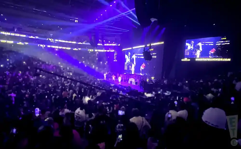 A concert crowd watches performers on a stage under bright purple lights, with screens showing live action.
