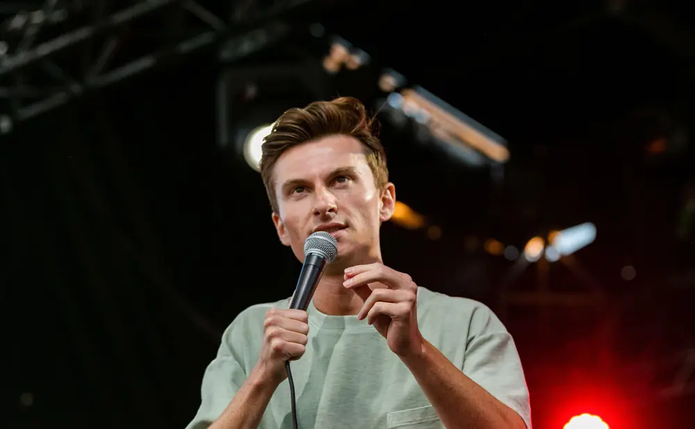 A man with light skin and brown hair holds a microphone and wears a light green t-shirt.