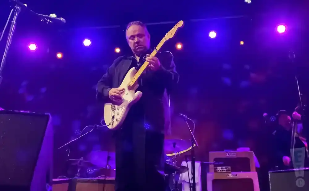 A man plays a white electric guitar on a stage with purple lights.