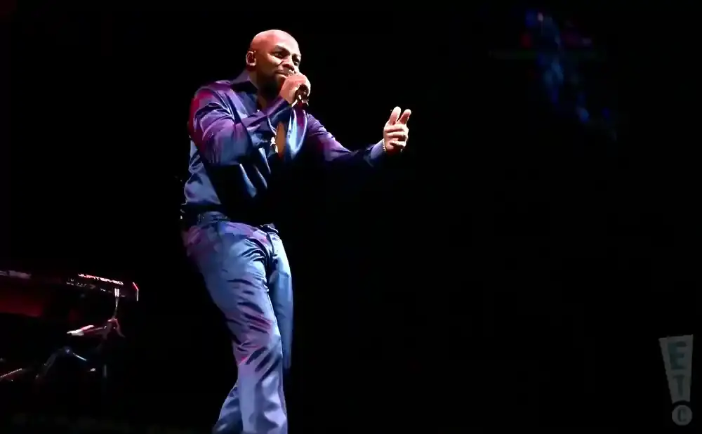 A bald Black man in a shiny blue shirt and pants sings into a microphone while gesturing with his free hand.