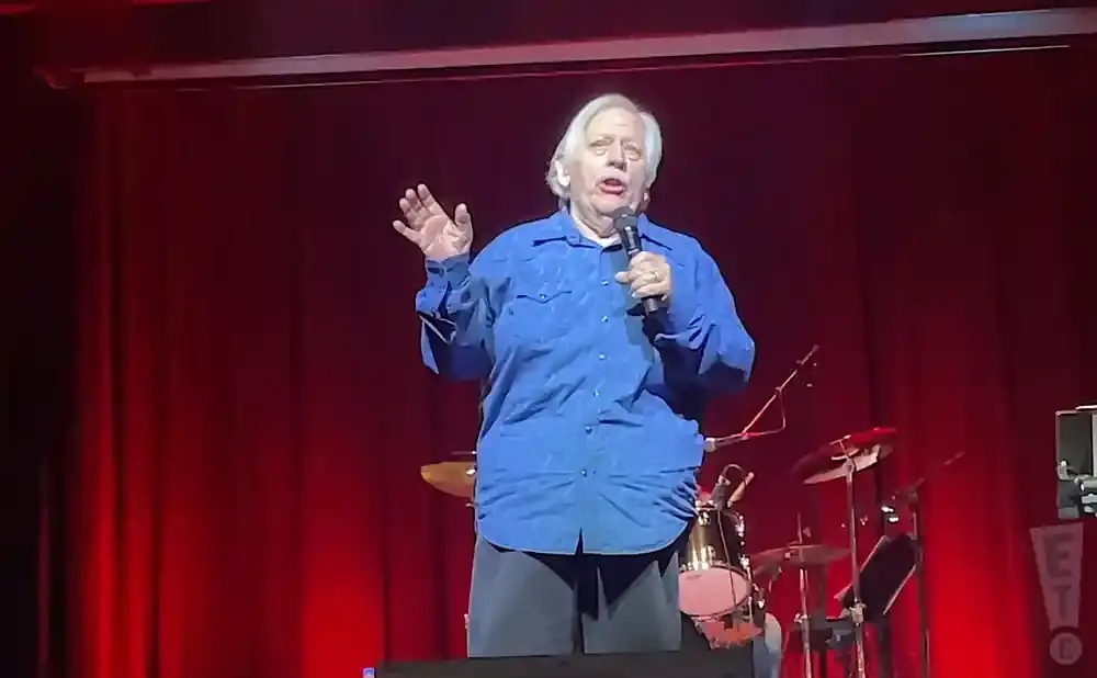A man in a blue shirt speaks into a microphone on a red stage.