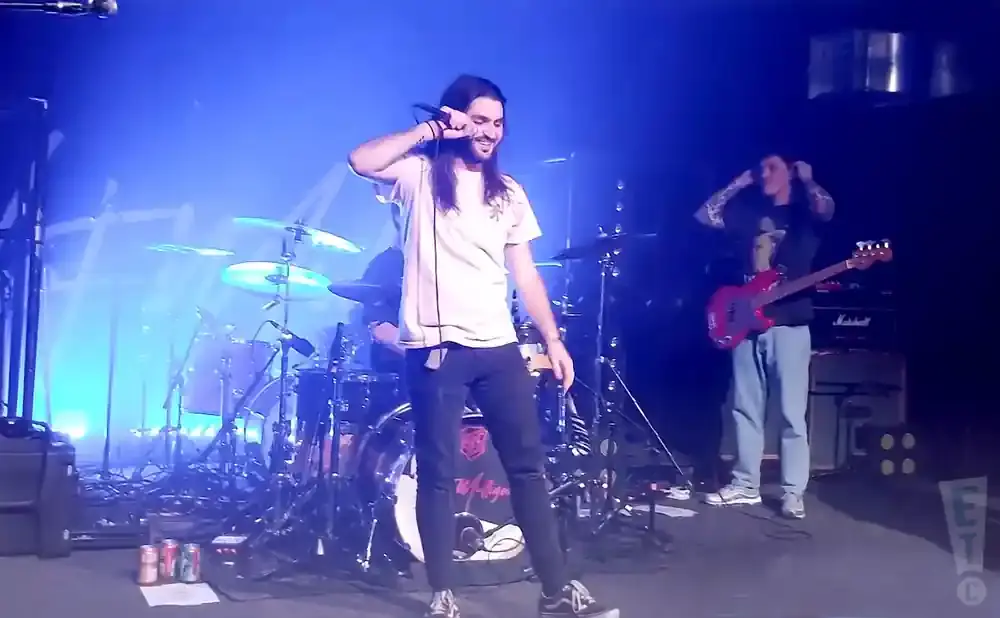 A male singer with long hair holds a microphone on stage, while another man plays a red bass guitar and a drummer is visible in the background.