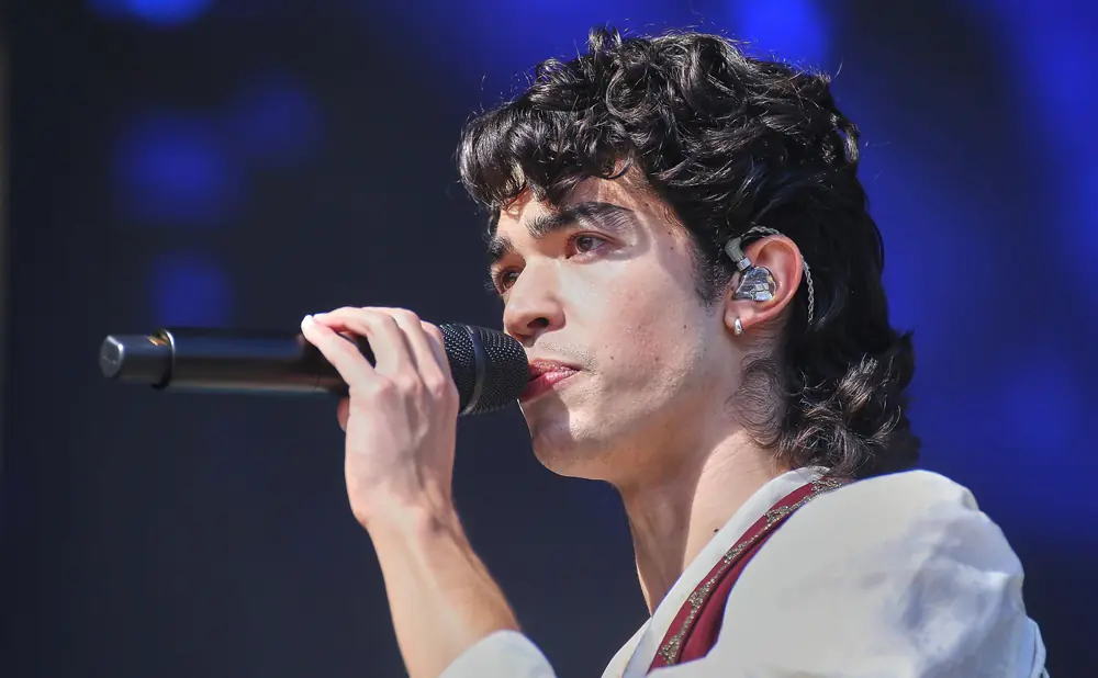 A male singer with curly dark hair holds a microphone to his lips while wearing an earpiece.
