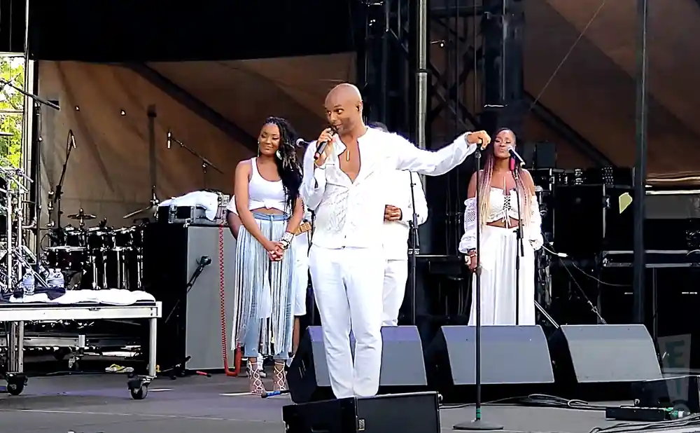A male singer in white addresses the audience while two women in white stand beside him on stage with drums in the background.