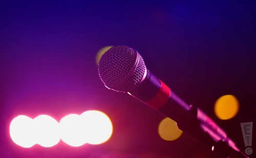 A microphone on a stand in front of purple and pink stage lights.