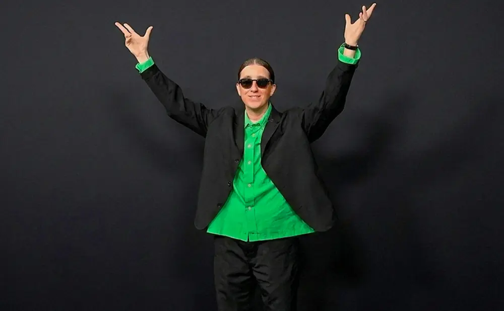 Person in a black suit and bright green shirt, arms raised, wearing sunglasses.