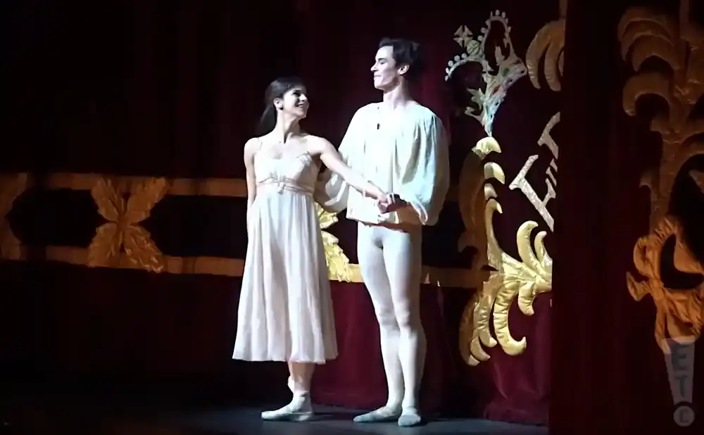 A ballet couple holds hands on stage; the woman wears a light dress, the man a light tunic and tights.