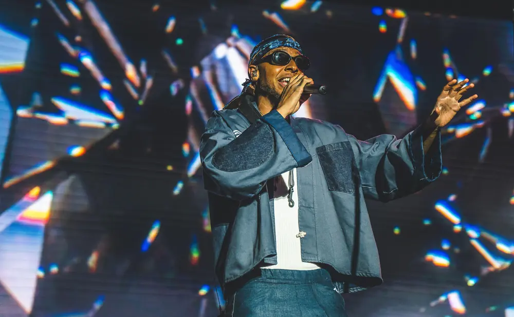 A man in sunglasses and a bandana performs on stage with a colorful, abstract background.