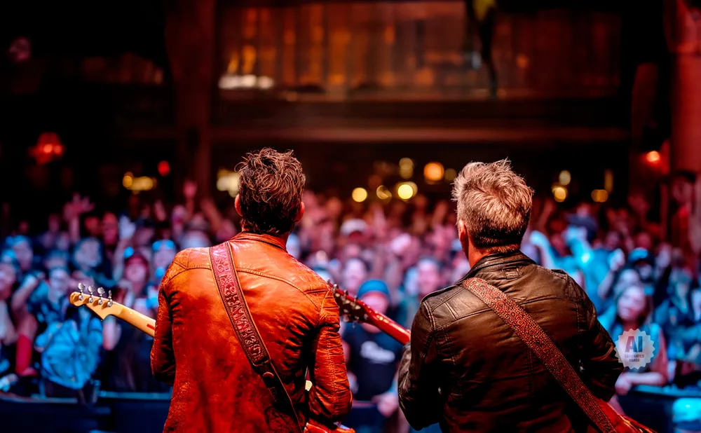 Two guitarists on stage face a cheering crowd, lit by stage lights.