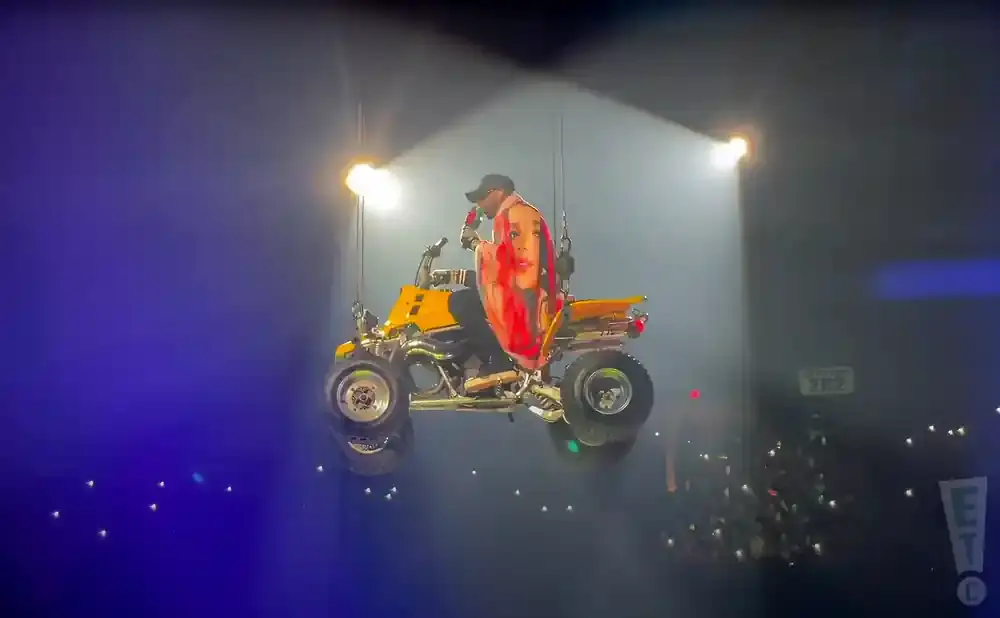 A performer on a yellow ATV is suspended in the air during a concert, holding a scarf with a woman's face on it.