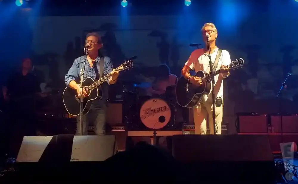 A band performs on stage, with two men in the foreground playing acoustic guitars and singing, and a drummer in the background.