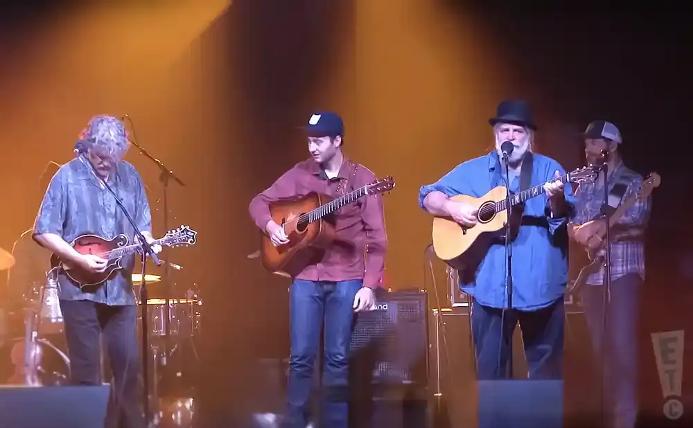A band plays on stage with warm lighting; one man plays a mandolin, two play guitars, and another plays a bass guitar.