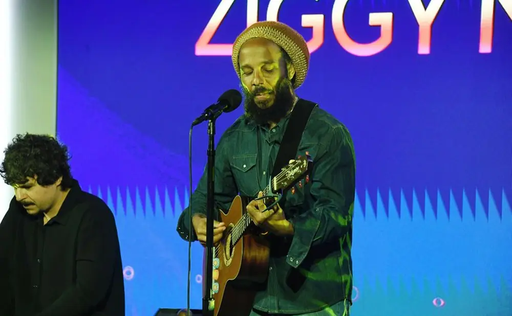 Ziggy Marley performs on stage with a guitar, wearing a beanie and denim shirt.