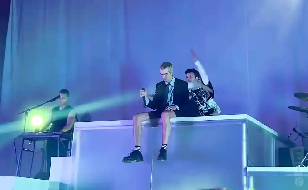 Singer in a suit and shorts sits on a platform holding a microphone, while another person behind him raises their arm.
