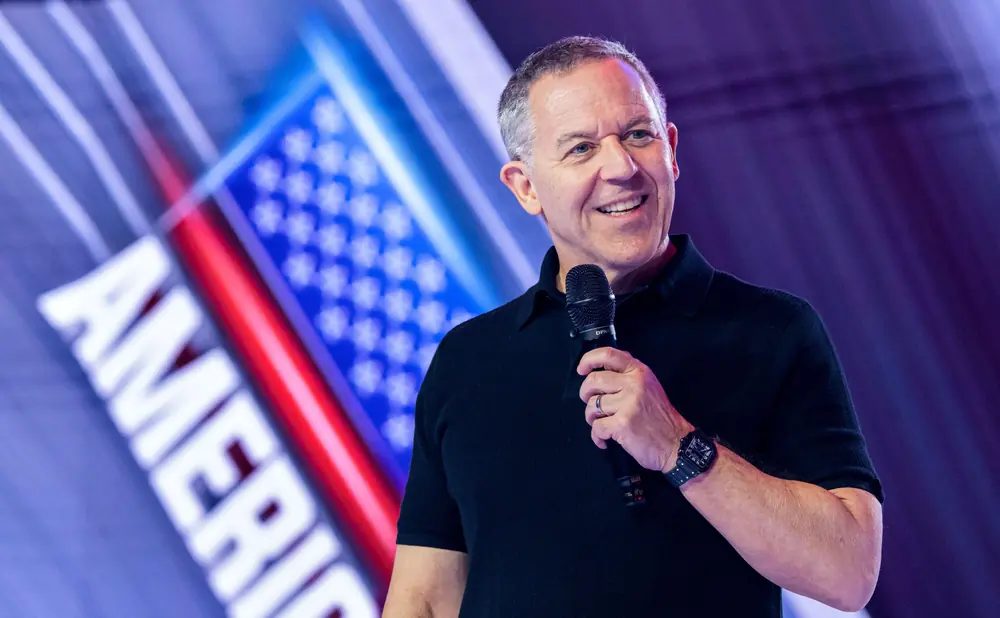 A man in a black shirt holds a microphone and smiles against a backdrop of the American flag.